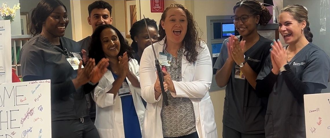 A woman in a lab coat smiles holding a large pair of ribbon-cutting scissors stands in the center as nursing students on either side smile and clap their hands
